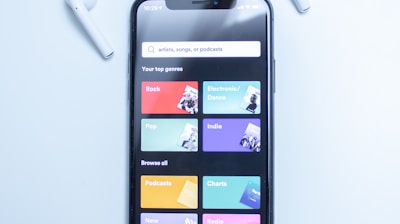 A smartphone displaying a music streaming app interface is placed on a light blue surface. The screen shows various musical genres and options in colorful boxes, including Rock, Electronic/Dance, Pop, Indie, Podcasts, Charts, New Releases, and Radio. Two white wireless earbuds are positioned on either side of the phone.