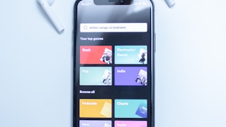 A smartphone displaying a music streaming app interface is placed on a light blue surface. The screen shows various musical genres and options in colorful boxes, including Rock, Electronic/Dance, Pop, Indie, Podcasts, Charts, New Releases, and Radio. Two white wireless earbuds are positioned on either side of the phone.
