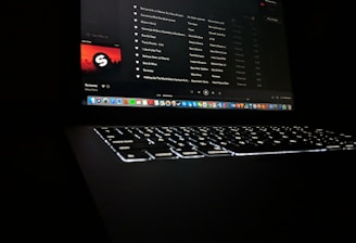 A darkly lit scene featuring a laptop with a glowing keyboard and a screen displaying a music streaming application. The interface shows a playlist with visible song titles and album artwork. Application icons are aligned at the bottom of the screen, indicating a computer interface.