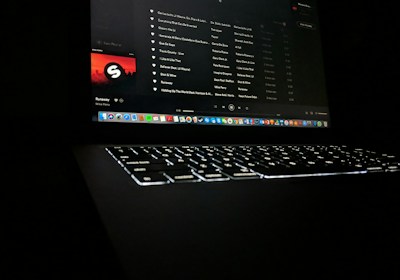 A darkly lit scene featuring a laptop with a glowing keyboard and a screen displaying a music streaming application. The interface shows a playlist with visible song titles and album artwork. Application icons are aligned at the bottom of the screen, indicating a computer interface.
