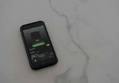 A smartphone with a dark case displays a music streaming app on the screen, placed on a light gray, smooth surface. The app interface is showing an album cover and music controls.
