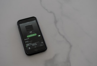 A smartphone with a dark case displays a music streaming app on the screen, placed on a light gray, smooth surface. The app interface is showing an album cover and music controls.