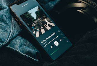 A smartphone displaying a music streaming application with an album cover visible on the screen. The phone is placed on denim fabric with a pair of black headphones partially visible alongside.