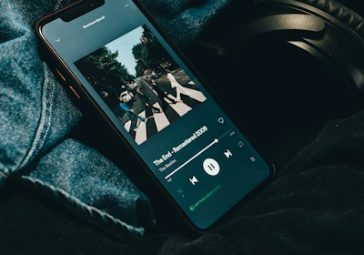 A smartphone displaying a music streaming application with an album cover visible on the screen. The phone is placed on denim fabric with a pair of black headphones partially visible alongside.