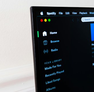 A computer screen displays the Spotify application, showing menu options such as Home, Browse, Radio, and Your Library, which includes Made For You, Recently Played, and Liked Songs. The application interface is sleek, with a dark background and white text, alongside green, yellow, and red window control buttons at the top left.