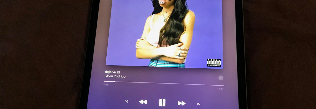 An electronic tablet displaying a music streaming application. The screen shows the cover art of a song, featuring a figure with long hair and a white top against a purple background. The person's face is decorated with colorful stickers. The music player interface is visible with playback controls and track information.