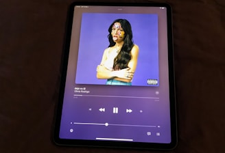 An electronic tablet displaying a music streaming application. The screen shows the cover art of a song, featuring a figure with long hair and a white top against a purple background. The person's face is decorated with colorful stickers. The music player interface is visible with playback controls and track information.