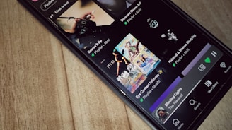 A smartphone is lying on a wooden surface, displaying a music streaming app that shows playlists and songs on the screen.