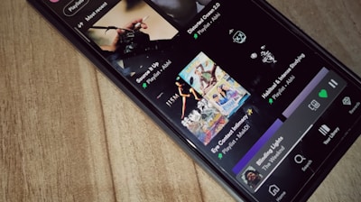 A smartphone is lying on a wooden surface, displaying a music streaming app that shows playlists and songs on the screen.